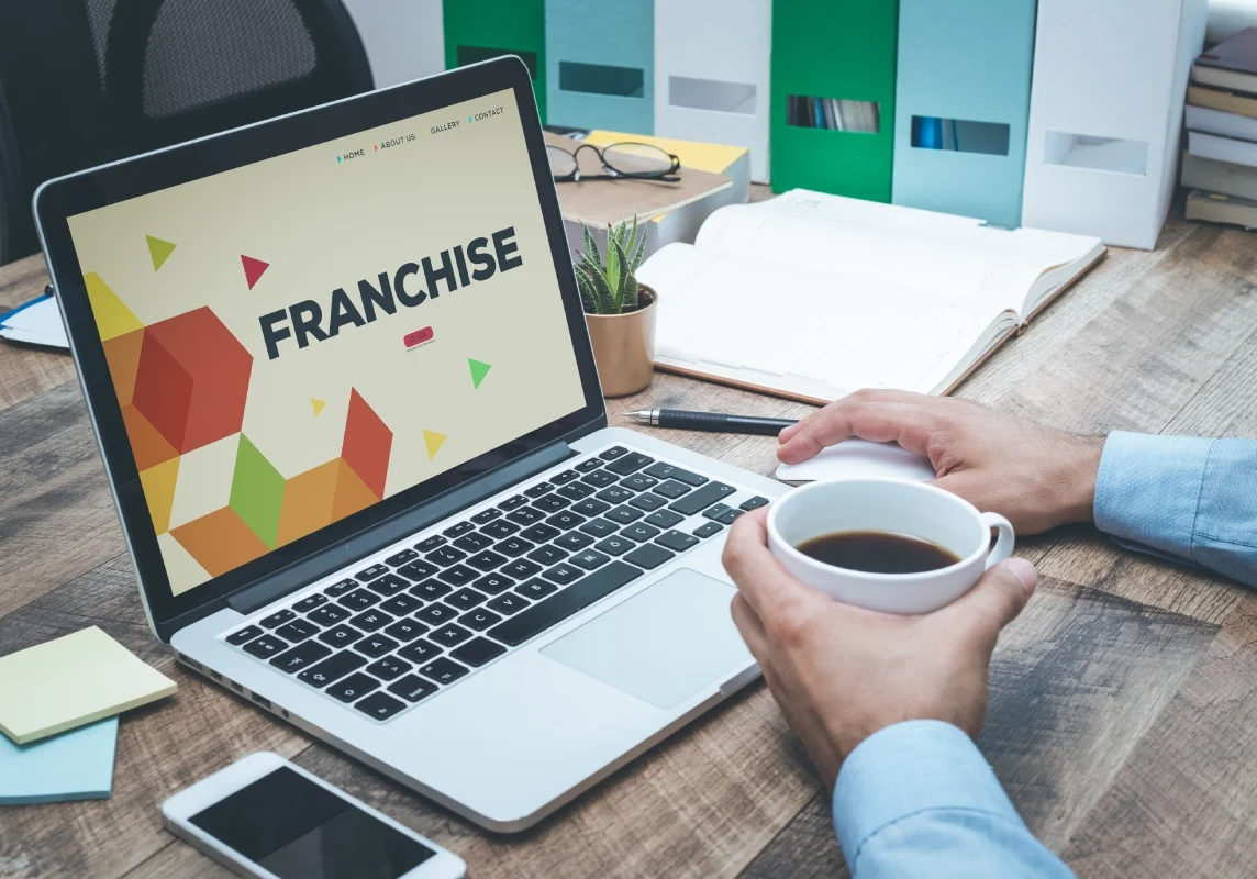 Laptop displaying "Franchise" with person holding coffee.